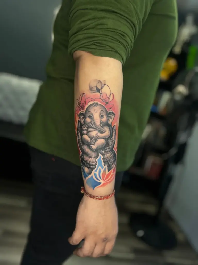 Spiritual baby Ganesha tattoo held in hands, with colour lotus and flowers, on the forearm