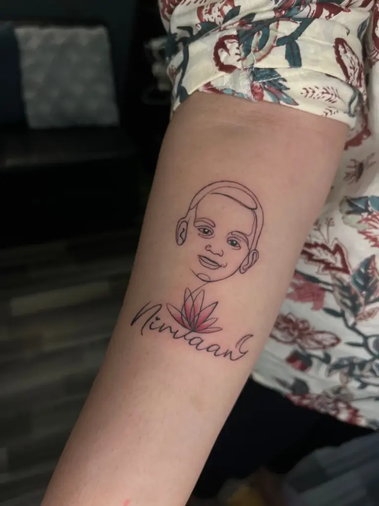 Single-line child portrait tattoo with name script and pink lotus on the forearm
